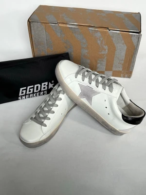 Vita GGDB sneakers med grå stjärna - Snygga vita sneakers från Golden Goose med grå stjärna på sidan, grå skosnören och svart detalj på hälen. Skorna har en klassisk låg siluett och är tillverkade i läder med slitna detaljer på sulan för en cool vintage-look.
