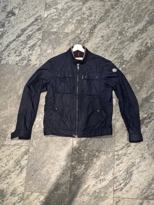 Moncler - Säljer min mörkblåa jacka från Moncler med klassisk logga på ärmen. Jackan har dragkedja framtill, flera fickor med knappare och en hög krage med röd-vit detalj. Perfekt till vår, höst och en frisk sommarkväll. Använd sparsamt. Storlek 3 motsv. S/M hör av er vid frågor!