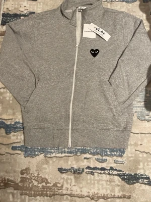 Grå zip hoodie Comme des Garçons - Snygg grå zip hoodie från Comme des Garçons PLAY med det ikoniska svarta hjärtat med ögon på bröstet. Tillverkad i mjuk bomullsmix och har ribbade muddar samt hög krage. Perfekt för en chill och stilren look.