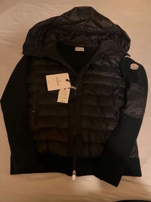 Svart pufferjacka från Moncler - Snygg svart pufferjacka från Moncler med huva och dragkedja framtill. NFC scannen på loggan fungerar(inga konstigheter). Jackan har stickade ärmar och klassisk Moncler-logga på ena ärmen. Perfekt för kyliga dagar och har en modern, sportig look. Inga skador, aldrig använd. Skriv om du har flera frågor mvh.