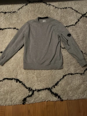 Grå sweatshirt från C.P. Company - Säljer en stilren grå sweatshirt från C.P. Company med rund halsringning och lång ärm. Tröjan har en ikonisk svart linsdetalj på ena ärmen och ribbade muddar vid ärmslut och nederkant. Perfekt för en avslappnad look. Pris går att diskuteras.