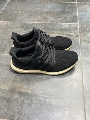 Ultra boost - Säljer ett par svarta Adidas Ultra Boost sneakers med stickad ovandel och klassiska tre ränder på sidan. Skorna har en vit Boost-sula som ger grym komfort och respons. Perfekta för dig som vill ha både stil och funktion i vardagen.