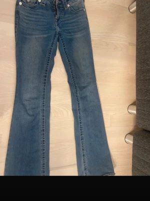 Blå bootcut jeans från True Religion - Säljer ett par klassiska blå bootcut jeans från True Religion med snygga kontrastsömmar och ikoniska ficklock bak. Jeansen har silverfärgade knappar och detaljerad brodyr på bakfickorna. Perfekta för dig som gillar en avslappnad men trendig look.