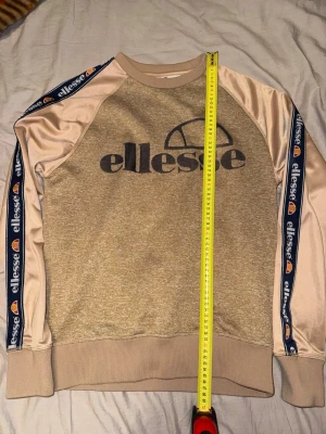 Beige sweatshirt från Ellesse - Snygg beige sweatshirt från Ellesse med stora loggan tryckt på bröstet. Tröjan har kontrasterande ärmar i ljusare beige och mörkblåa band med Ellesse-logga längs ärmarna. Ribbstickade muddar vid ärmslut och nederkant. Perfekt för en sportig och avslappnad stil.