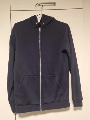 Mörkblå zip hoodie med fickor - En enkel och stilren mörkblå hoodie med dragkedja framtill och två stora fickor. Hoodien har huva med snörning och ribbade muddar vid ärmslut och nederkant. Perfekt för en avslappnad look. Materialet känns mjukt och bekvämt, troligen bomullsblandning.