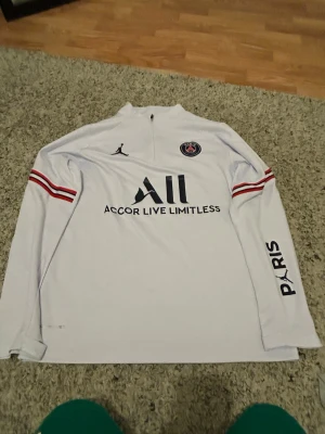 Vit PSG träningströja Jordan - Säljer en vit långärmad träningströja från PSG x Jordan med halv dragkedja. Tröjan har svarta och röda detaljer, PSG-logga på bröstet, Jordan-logga och texten 'Accor Live Limitless' framtill samt 'PARIS' på ärmen. Perfekt för fotbollsträning eller sport.