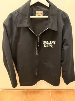 Svart jacka från Gallery Dept. - Stilren svart jacka från Gallery Dept. med vit logga tryckt på bröstet. Jackan har klassisk krage, dragkedja framtill och två sidofickor. Tillverkad i bomull och har en normal passform som passar till många olika looks.