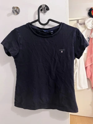 Mörkblå t-shirt från GANT - Snygg mörkblå t-shirt från GANT med klassisk passform och rund hals. T-shirten har korta ärmar och en liten broderad GANT-logga på bröstet. Perfekt basplagg i mjuk bomull som funkar till allt. Storlek 158-164