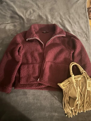 Vinröd teddyjacka med dragkedja - Mysig vinröd teddyjacka med dragkedja framtill och stora fickor. Jackan har en hög krage och är tillverkad i fluffigt teddy-material som ger en avslappnad och trendig look. Perfekt för kyliga dagar och enkel att matcha med olika outfits.