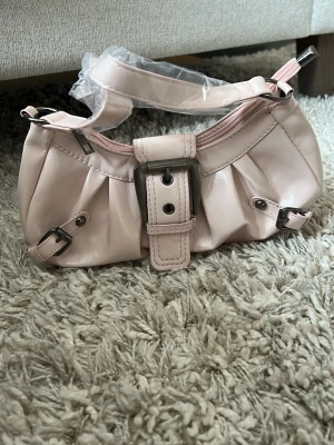 Ljusrosa axelremsväska med spänne - Söt ljusrosa handväska i glansigt konstläder med silverfärgade detaljer och stora dekorativa spännen framtill. Väskan har en kort axelrem och dragkedja upptill. Perfekt för dig som gillar trendiga accessoarer med en touch av Y2K-stil. Ej äkta läder (plast-ish material) notera att dragkedjan kan vara lite trög att dra igen.