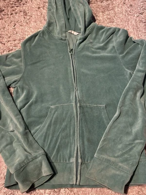 Grön hoodie med dragkedja - Mysig grön hoodie i mjuk velour med dragkedja framtill och kängurufickor. Klassisk huva och långa ärmar. Perfekt för en avslappnad och chill stil. Passar dig som gillar bekväma och trendiga plagg.