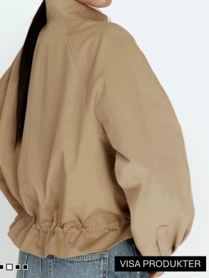 Slutsåld oanvänd jacka i bomullstwill med lappen kvar  - Stilren beige vindjacka med hög krage och dragsko i midjan för en snygg, avslappnad passform. Jackan har två fickor framtill med knappstängning och raka ärmar. Perfekt för lager-på-lager och enkel att  matcha till jeans.