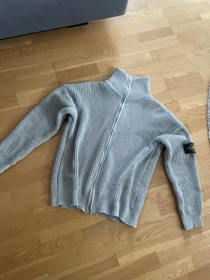 Ljusgrå stickad tröja Stone Island - Säljer en ljusgrå stickad tröja från Stone Island med hög krage och ribbstickad struktur. Tröjan har lång ärm och den klassiska Stone Island-loggan på vänster ärm. Perfekt för dig som gillar streetwear och snygga detaljer. M men passar S också, helt ny oanvänd 
