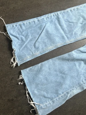 Ljusblåa vida jeans med rå kant - Säljer ett par ljusblå jeans med riktigt breda ben och råa, fransiga benslut. Klassisk femficksmodell i denim med hög midja. Perfekta för dig som gillar en avslappnad och trendig stil. Materialet är 100% bomull för en skön känsla.