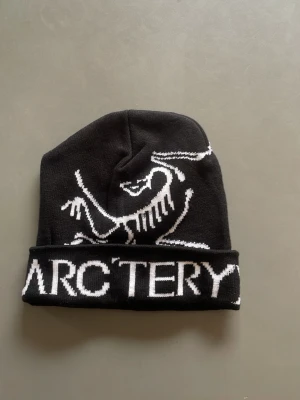 Svart mössa från Arc'teryx - Snygg svart mössa från Arc'teryx med vit logga och text framtill. Mössan har uppvikt kant och är tillverkad i mjukt, stickat material som sitter skönt på huvudet. Perfekt för dig som gillar outdoor och streetwear vibes.