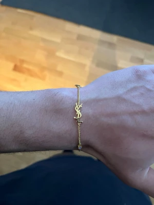 Guldigt armband från Saint Laurent - Snyggt och stilrent armband i guldton från Saint Laurent med den ikoniska YSL-loggan framtill. Kedjan är tunn och justerbar med ett litet hjärta och en bricka med märkets namn på baksidan. Perfekt accessoar för dig som gillar exklusiv och trendig design.