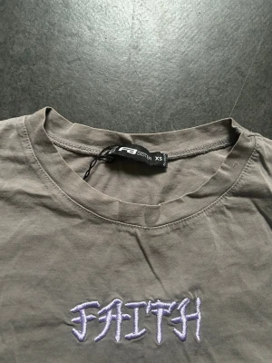 Grå t-shirt med broderad text FAITH - Snygg grå t-shirt från FB Sister i storlek XS. T-shirten har en rak passform och är tillverkad i mjuk bomull. På bröstet finns broderad texten 'FAITH' i vitt och lila, vilket ger en cool detalj till looken.