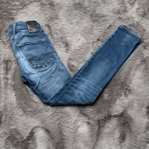 Blå jeans från Replay - Säljer ett par blå Replay anbass jeans med klassisk femficksdesign och snygga slitningar framtill. Jeansen har en skinnpatch med logga bak i midjan och raka ben. Perfekta för en avslappnad och trendig look. W27 L32. Priset är ej hugget i sten, hör av er vid minsta fundering!✨