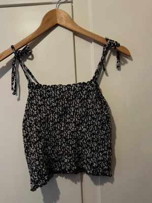 Blommig svart linnetopp med knytband - Söt svart linnetopp med små vita blommor och knytbara axelband. Toppen har smockad resår upptill och är croppad i modellen, perfekt för varma dagar. Passar dig som gillar en avslappnad och trendig stil.