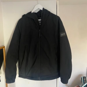 Svart vindjacka från Calvin Klein - Svart vindjacka från Calvin Klein med huva och dragkedja framtill. Jackan har två sidofickor med dragkedja och en diskret logotyp på ärmen. Tillverkad av 100% återvunnen nylon, perfekt för dig som gillar clean och enkel stil.