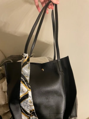 Svart shoppingväska med scarfdetalj - Stilren svart shoppingväska i syntetmaterial med långa handtag och en snygg scarfdetalj i svart, vitt och guld. Väskan har enkel knappstängning upptill och rymligt huvudfack, perfekt för att bära allt du behöver till skolan eller stan.