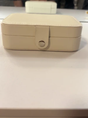 Beige smyckesask i läderimitation - Stilren smyckesask i beige läderimitation med rundade hörn och tryckknapp framtill. Perfekt för att förvara ringar, örhängen och halsband. Kompakt och enkel att ta med på resan eller ha på sminkbordet.