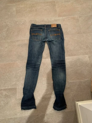  Nudie Jeans - Säljer ett par blå slim fit jeans från Nudie Jeans med slitningar och snygga detaljer på bakfickorna. Dom är waist 27 men sitter som 28/29 Jeansen har klassisk femficksmodell, låg midja och råa kanter vid bensluten. Tillverkade i denim med cool tvätt och kontrastsömmar.