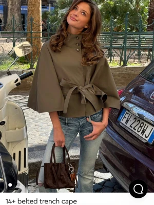 Belted trench cape - Stilren olivgrön trenchcape från gina med knappar framtill och knytskärp i midjan. Capen har vida ärmar och hög krage för en modern look. Perfekt att slänga över en outfit för extra edge. 