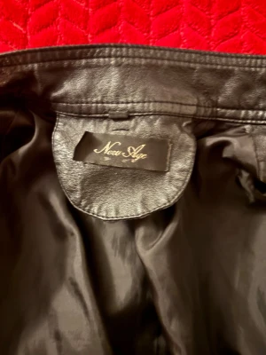 Svart skinnjacka från New Age - Snygg svart skinnjacka från New Age med flera dragkedjeförsedda fickor och ståkrage med tryckknapp. Jackan har en klassisk bikerstil och detaljerade sömmar som ger en cool look. Perfekt för dig som vill ha en stilren och trendig jacka.