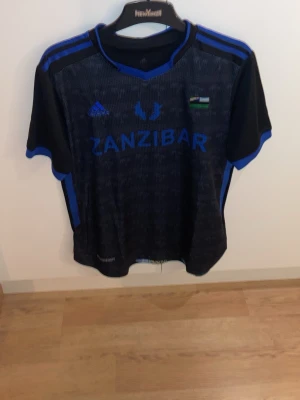 Svart Zanzibar fotbollströja Adidas - Säljer en svart fotbollströja från Adidas med blå detaljer och texten 'ZANZIBAR' på bröstet. Tröjan har korta ärmar, rund hals och en liten flagga på bröstet. Perfekt för dig som gillar unika matchtröjor och sportig stil.