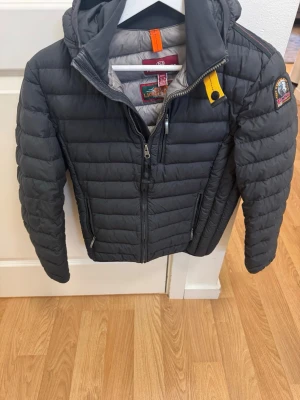 Mörkgrå svart dunjacka från Parajumpers - Snygg mörkgrå dunjacka från Parajumpers med huva, gul detalj vid halsen och patch på ärmen. Jackan har två sidofickor med dragkedja och quiltad design. Perfekt för kalla dagar och riktigt trendig look.stl Y-S motsvarar typ en xs 