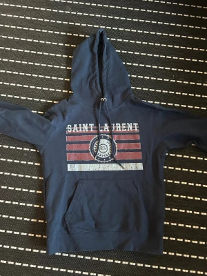 Blå Saint Laurent hoodie med tryck - Mörkblå hoodie från Saint Laurent med stor huva och dragsko. Framsidan har ett coolt tryck med texten 'SAINT LAURENT' och amerikanskinspirerade ränder i rött och vitt. Stor magficka framtill och ribbade muddar. Perfekt för en avslappnad streetstyle-look.
