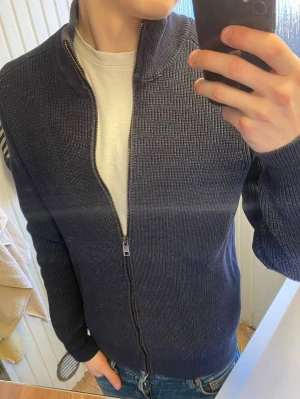 Mörkblå ziptröja från MNG Man - Säljer en mörkblå stickad ziptröja från MNG Man. Tröjan har hög krage, hel dragkedja framtill och ribbstickad struktur. Perfekt att slänga på sig över en t-shirt för en clean och enkel look.