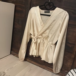 Champagnefärgad omlottblus Missäy - Snygg champagnefärgad omlottblus från Missäy i glansigt satintyg. Blusen har långa ärmar, knytband i midjan och volang nedtill. Andvänt fåtal gånger. Original pris 599.