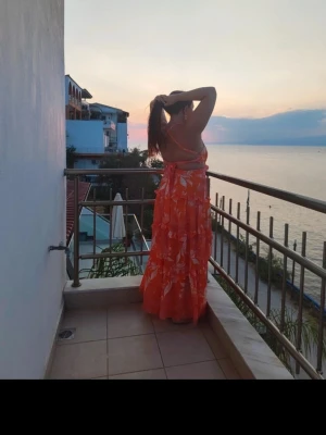 Orange långklänning med volanger - Säljer en färgstark orange långklänning med vita blommönster och volangdetaljer. Klänningen har smala axelband och öppen rygg med knytning. Perfekt för varma sommardagar och har en luftig passform.