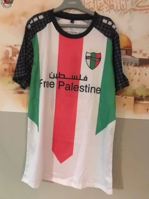Palestino T-shirt med tryck/ 3 xl  - Storlek xxxl (3XL). Unik T-shirt från Palestino med tryck 'Free Palestine' och klubbens emblem på bröstet. Tröjan har korta ärmar i svartmönstrat tyg, samt stora fält i vitt, rött och grönt framtill. Baksidan har svart grafik och text på flera språk. Perfekt för dig som vill visa ditt stöd.