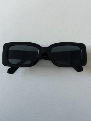 Svarta rektangulära solglasögon - Stilrena svarta solglasögon med rektangulära bågar och mörka glas. Snygg och modern design som passar perfekt till en trendig look. Bågarna är i plast och ger en cool vibe till din outfit.