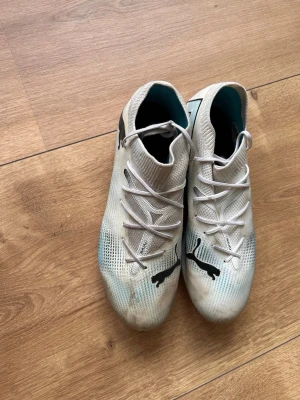 Vita Puma fotbollsskor med rosa sula - Säljer ett par vita Puma fotbollsskor med svart logga och coola grafiska detaljer på sidan. Skorna har snörning, låg profil och en grym rosa sula med dobbar för bra grepp på planen. Ovandelen är i syntetmaterial och insidan är turkos. Perfekta för dig som vill sticka ut på matchen!