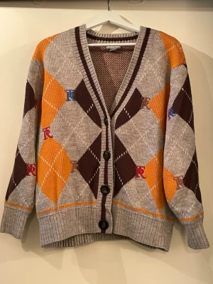 Argylemönstrad kofta med knappar - Kofta med klassiskt argylemönster i beige, brunt och orange samt broderade bokstäver i rött, blått och gult. Djup V-ringning, ribbade muddar och tre stora knappar framtill. Mjuk och bekväm. 