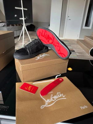 Svarta sneakers från Christian Louboutin - Stilrena svarta sneakers i mocka från Christian Louboutin med den ikoniska röda sulan och diskret logga på sidan. Skorna har svarta skosnören, lågt skaft och klassisk siluett. Perfekta för dig som vill ha en exklusiv touch på din outfit.