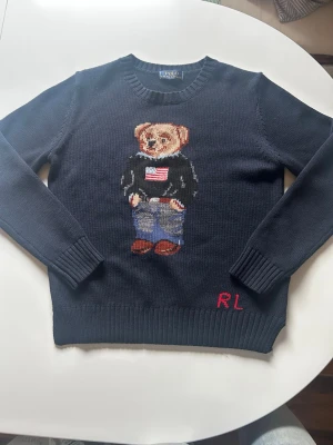 Mörkblå stickad tröja Polo Bear Ralph lauren - Mörkblå stickad tröja från Ralph Lauren med den ikoniska Polo Bear på bröstet. Tröjan har rund hals, långa ärmar och ribbade muddar. RL-broderi i rött nertill. Tröjan är i storlek L men jag skulle säga att den passar dig som har xs/s. Jag är 172 och säljer då den är för kort i armarna för mig.