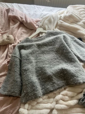 Grå stickad tröja från Zara - Mysig grå stickad tröja från Zara med boxy passform och trekvartsärmar. Tröjan har en rund halsringning och är tillverkad i ett fluffigt, mjukt material som ger en avslappnad look. Perfekt att matcha med jeans eller kjol.