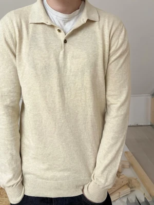 Beige stickad tröja med krage - Snygg beige stickad tröja från John Henric gjord av merinoull, med enklassisk krage och knappar vid halsen. Tröjan har långa ärmar och ribbade muddar vid ärmslut och nederkant. Perfekt för lager på lager och enkel att matcha med jeans. Modellen är 185cm och tröjan är lite för liten.