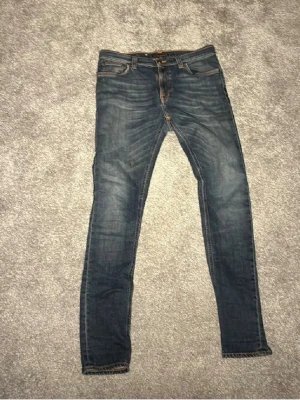 Nudie jeans - Säljer ett par mörkblå slim jeans med snygga och tvättade detaljer. Klassisk femficksmodell med dragkedja och knapp. Jeansen har kontrastsömmar och är i stretchigt material för skön passform.