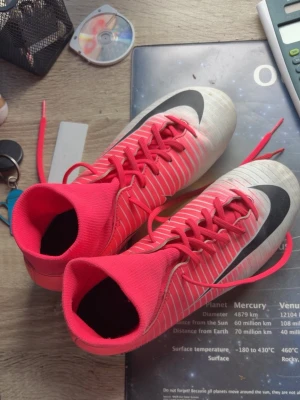 Nike Mercurial rosa fotbollsskor - Säljer ett par Nike Mercurial fotbollsskor i rosa och vitt med svarta detaljer. Skorna har hög strumpkrage för extra stöd, snörning och klassisk Nike-logga på sidan. Perfekta för dig som vill sticka ut på planen och få bra grepp tack vare dobbarna.
