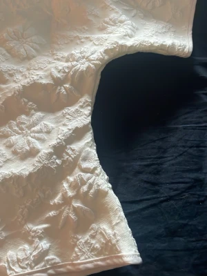 Vit blommig blus med struktur - Supersöt vit blus med korta ärmar och upphöjt blommönster i tyget. Blusen har en boxig passform och känns både trendig och unik tack vare den strukturerade ytan. Perfekt att styla med jeans eller kjol för en fräsch look.