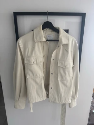 Krämvit overshirt från Bershka - Stilren krämvit overshirt från Bershka med två stora bröstfickor med lock och tryckknappar. Rak passform, klassisk krage och bälte i midjan för extra detalj. Tillverkad i ett mjukt bomullsliknande material. Perfekt för lager på lager och trendig look.