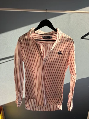 Randig skjorta från Polo Ralph Lauren - Snygg skjorta från Polo Ralph Lauren i vitt med röda och blå ränder. Klassisk krage, långärmad och med broderad logga på bröstet. Perfekt för dig som gillar en stilren och tidlös look. Aldrig använt den och priset är prutbart 