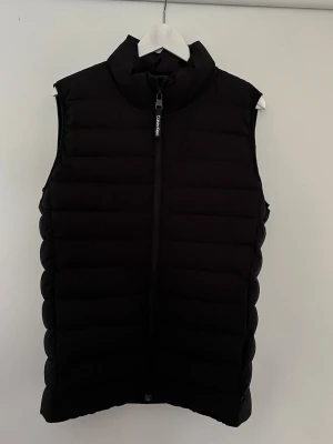 Dunväst från Calvin Klein - Snygg svart dunväst från Calvin Klein med hög krage och dragkedja framtill. Västen har en stilren design med horisontella sömmar och diskret logga på dragkedjan. 3 månader gammal, använd typ 3 gånger. Hör av dig vid frågor. 