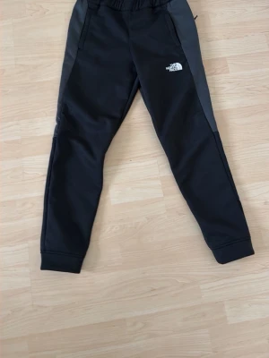 Svarta mjukisbyxor The North Face - Snygga svarta mjukisbyxor från The North Face med grå sidopaneler och vit logga på låret. Byxorna har resår i midjan, två fickor och muddar vid bensluten. Perfekta för chill eller träning, riktigt sköna och stilrena.